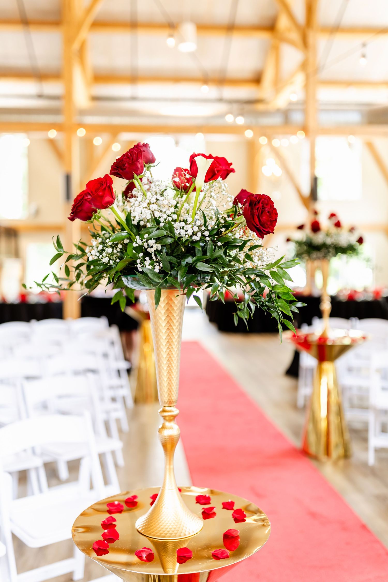 Red roses and baby's breath arranged in gold pedestal vase along red carpet aisle.