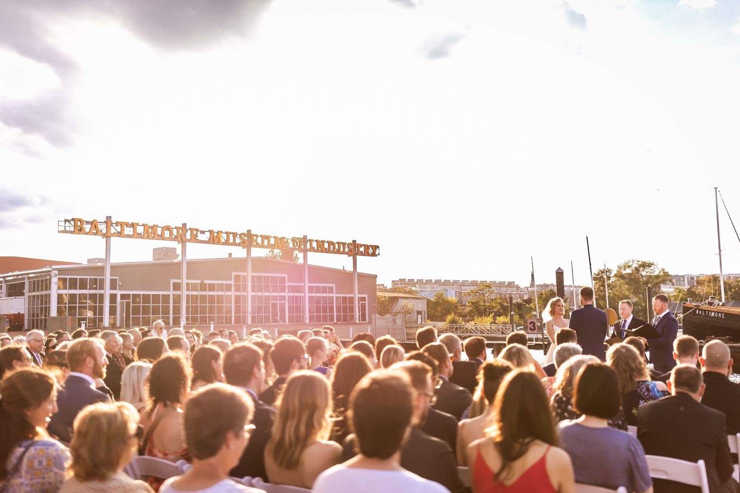 Large crowd gathers at an outdoor venue during golden hour with sunlight creating a warm atmospheric glow.