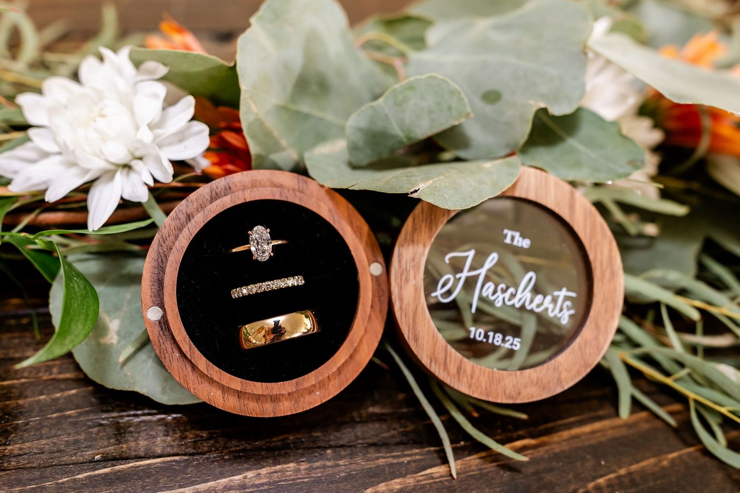 Wedding rings displayed in wooden ring boxes with eucalyptus leaves and floral decorations on rustic surface.