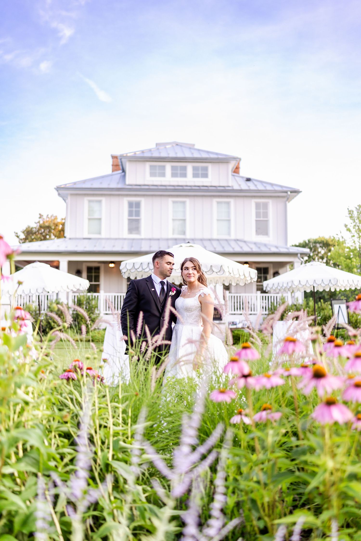 A white colonial-style house with wraparound porch serves as backdrop to a garden filled with pink cosmos flowers.