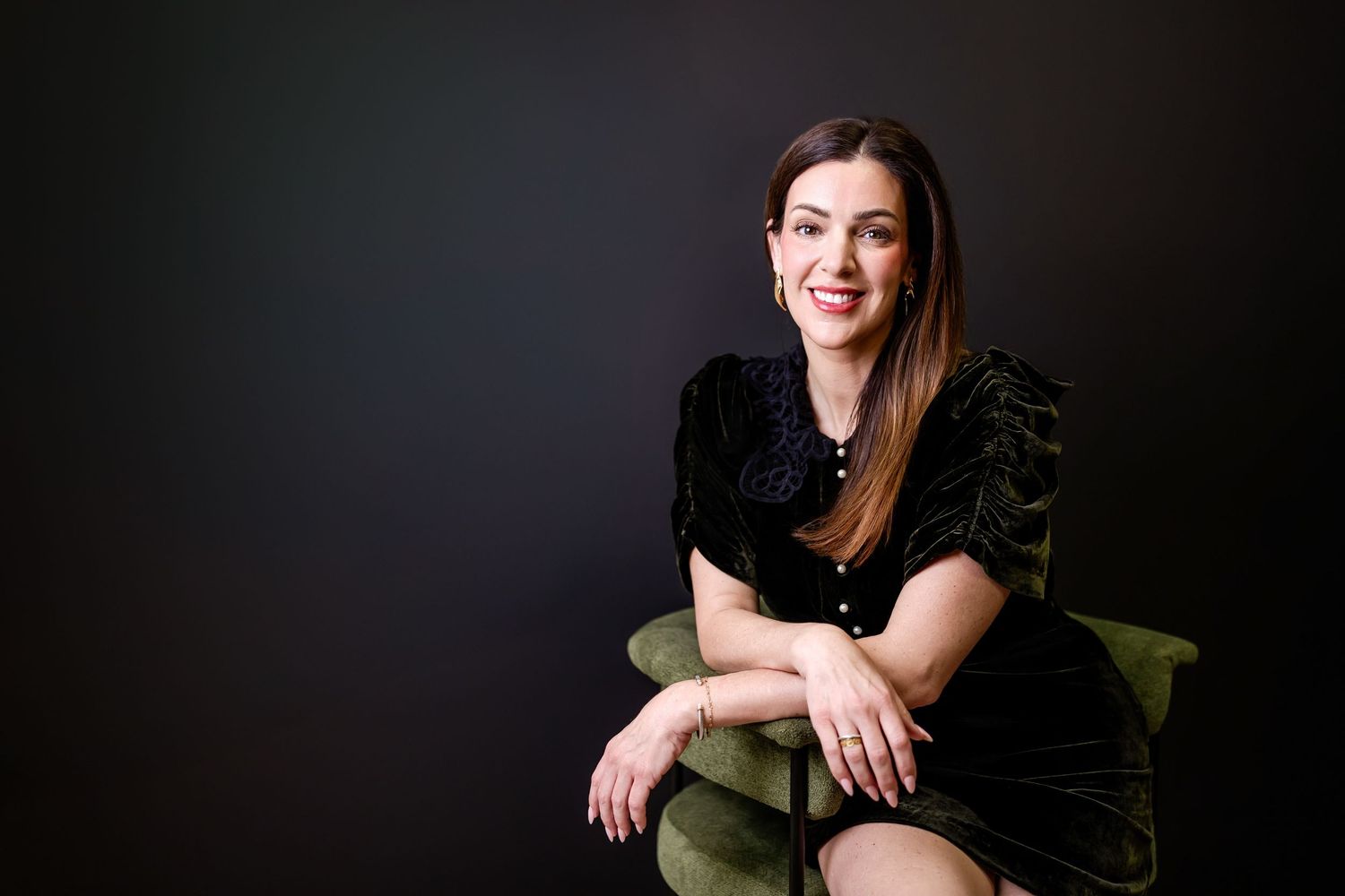 Woman in black velvet dress sitting gracefully in a green chair, smiling confidently against a dark studio background.