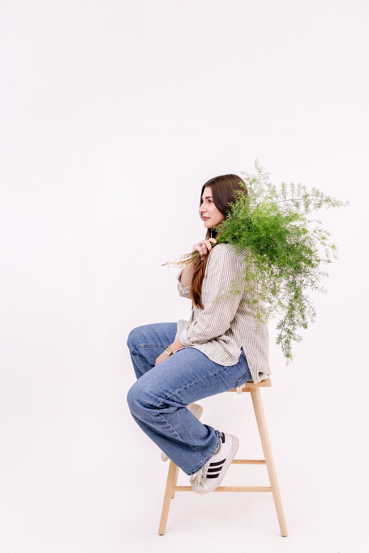 Young woman sitting on wooden chair holding white wildflowers, wearing jeans and striped shirt, white background.