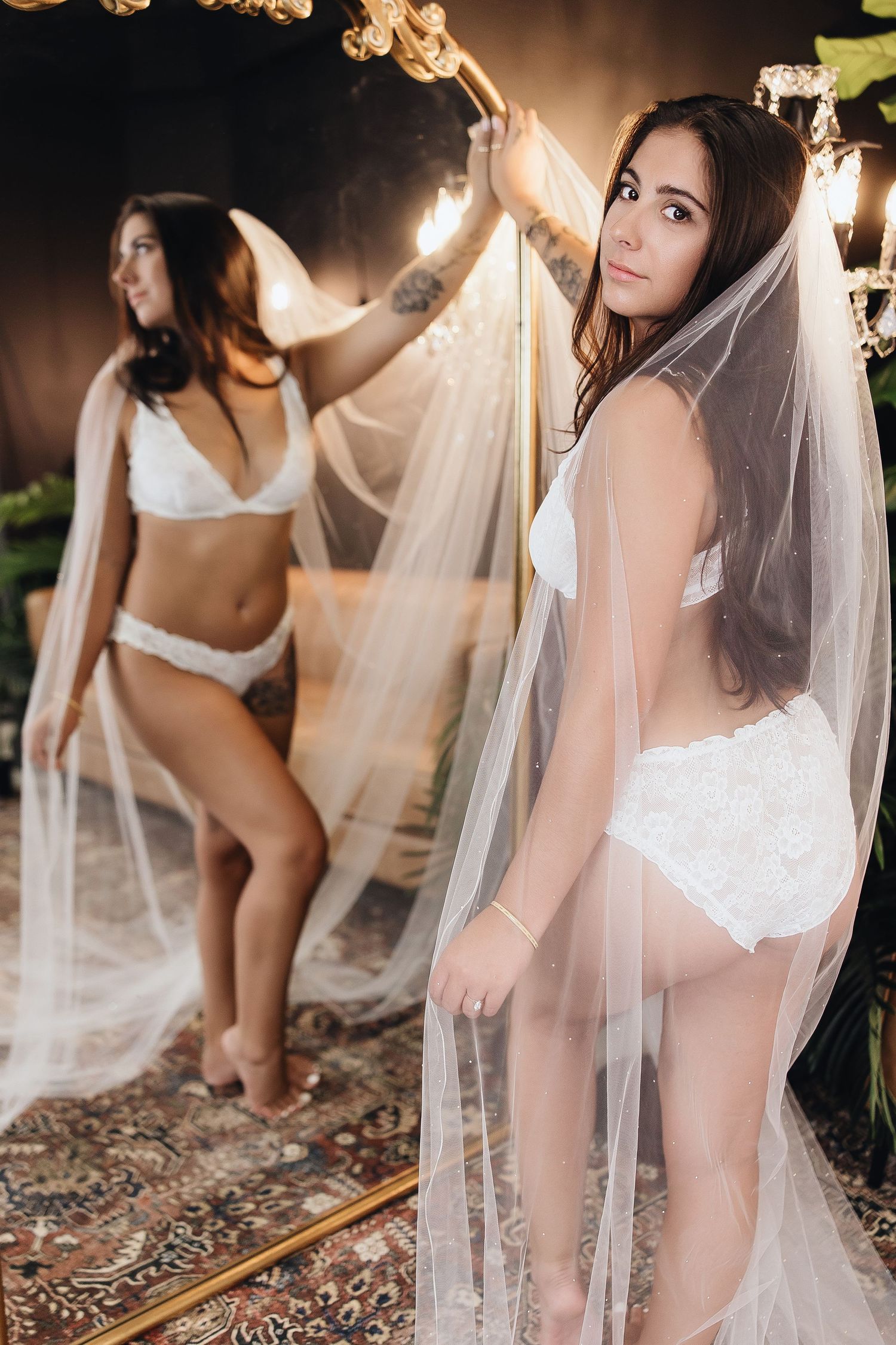 Bride in white lace lingerie and veil posing near mirror in elegant room with warm lighting.