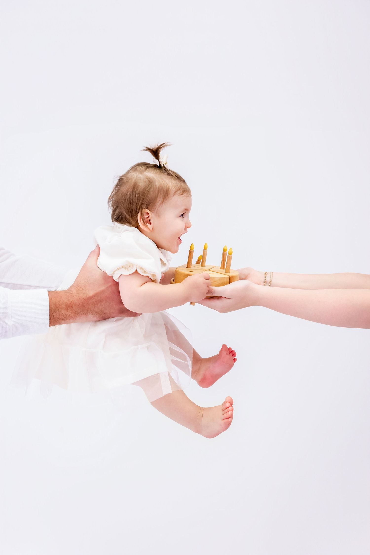 Baby in white dress reaching for a golden crown being passed between adult hands against white background.