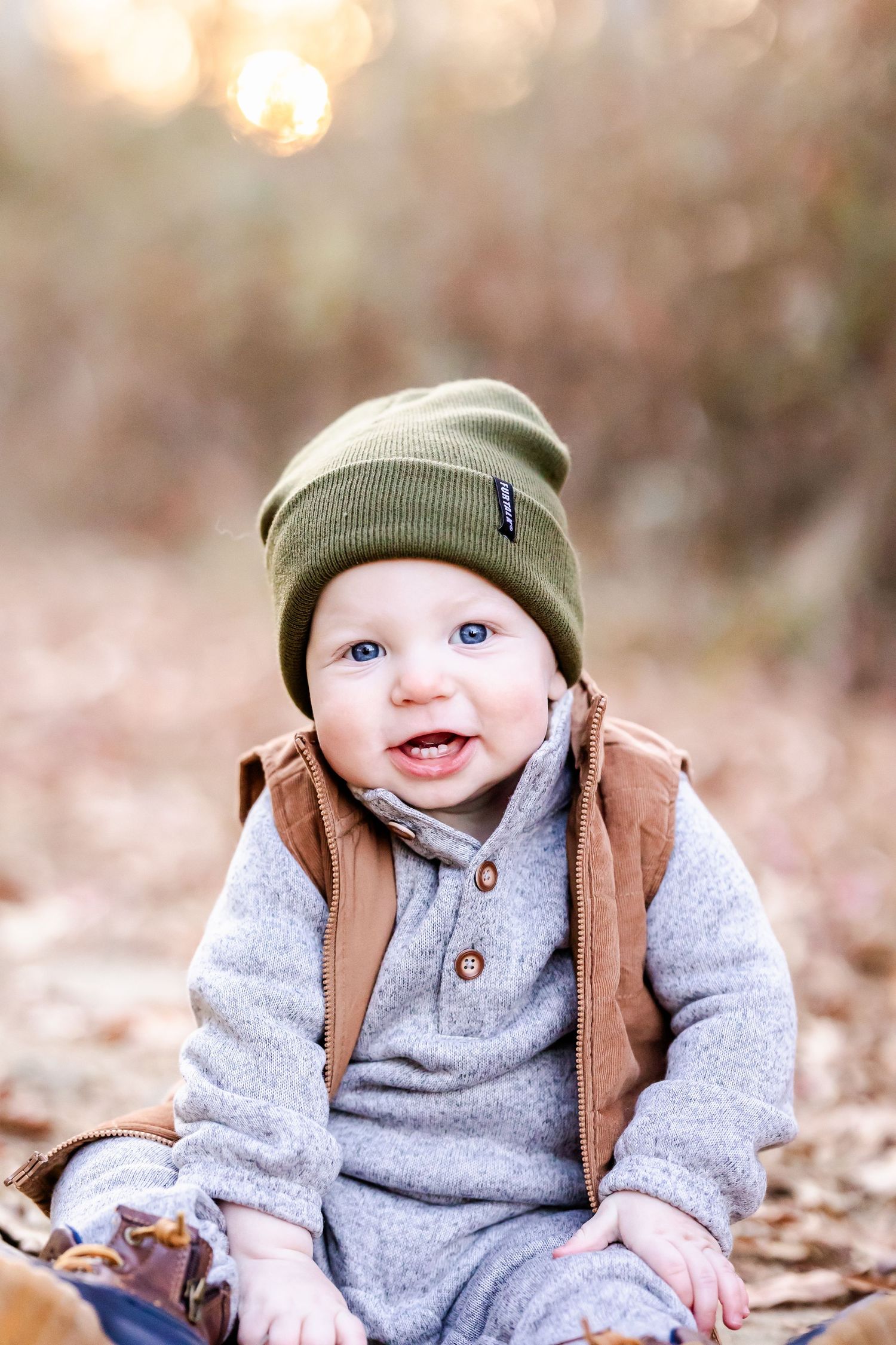 A baby wearing a green beanie hat and gray sweater sits on fallen autumn leaves with a brown backpack.