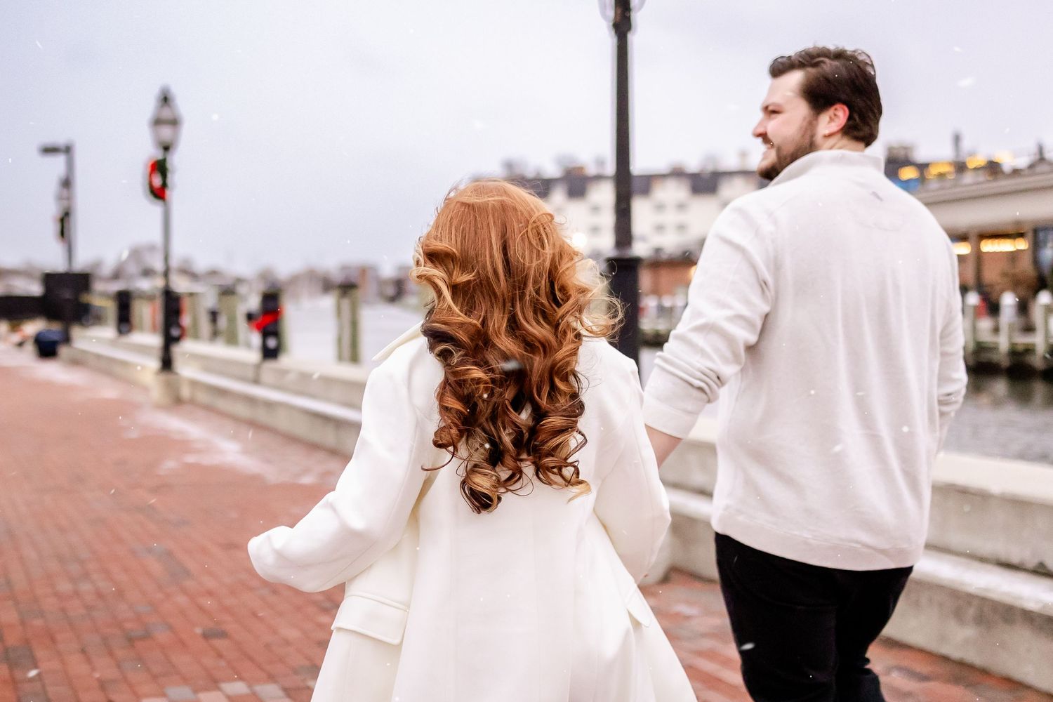 A couple in white winter coats walking along a brick waterfront promenade on an overcast day.