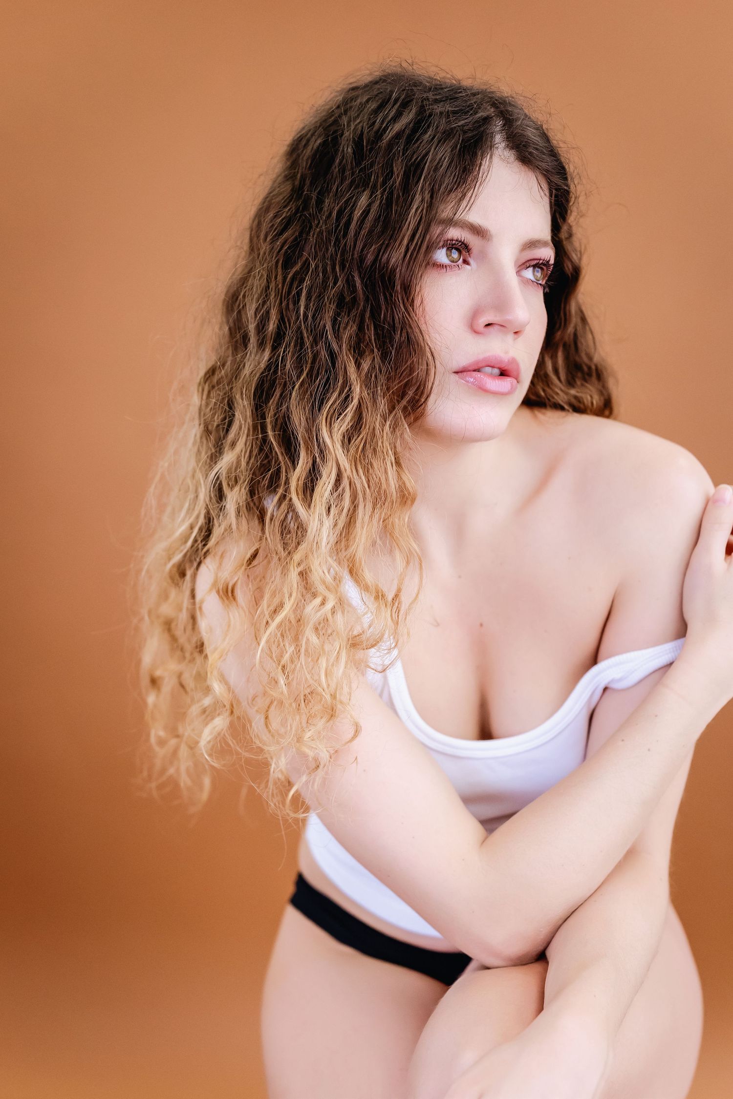 Person with ombre curly hair wearing white top poses against warm brown background in artistic portrait series.