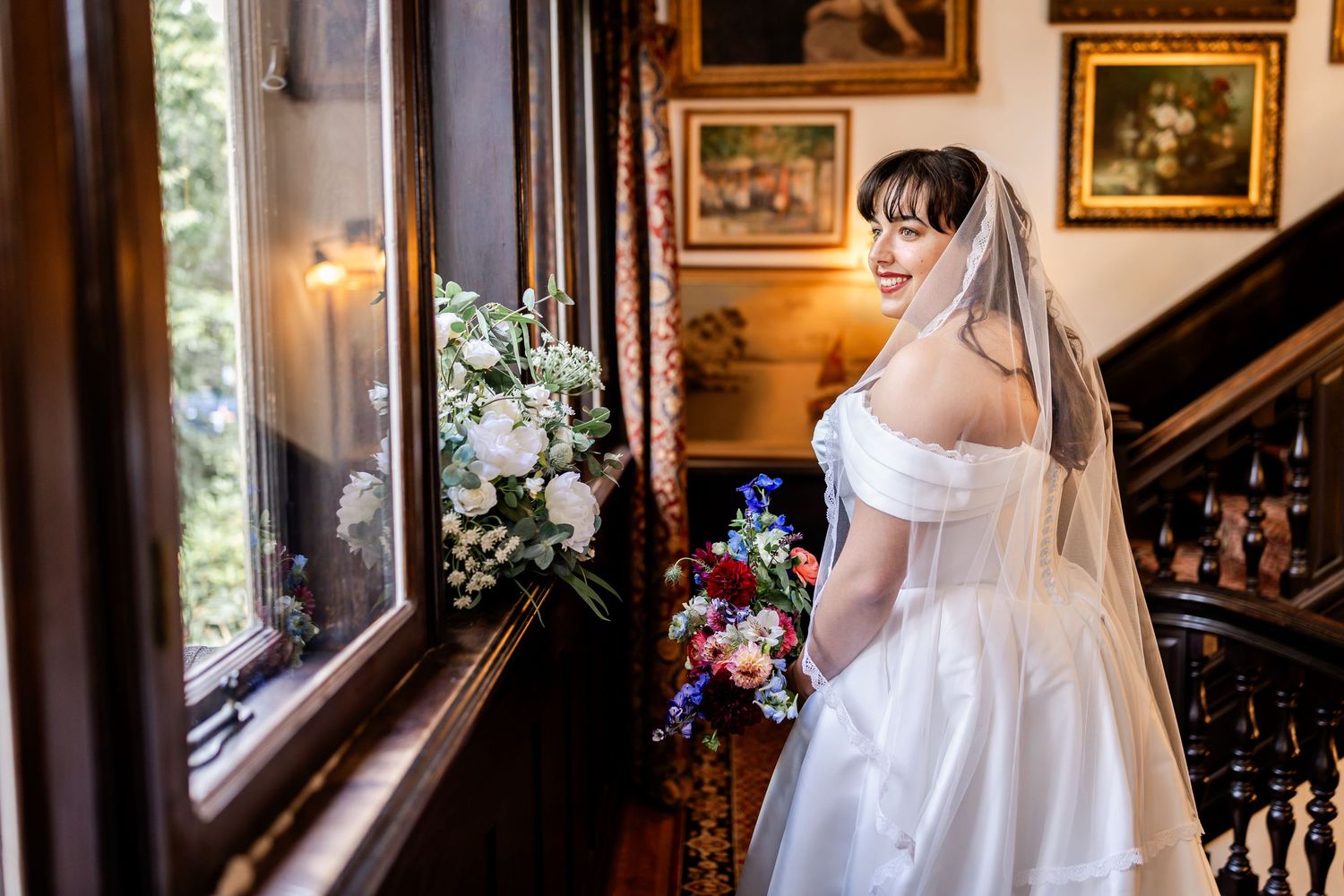 Bride in off-shoulder white gown and veil holds bouquet by ornate wooden staircase in elegant venue.