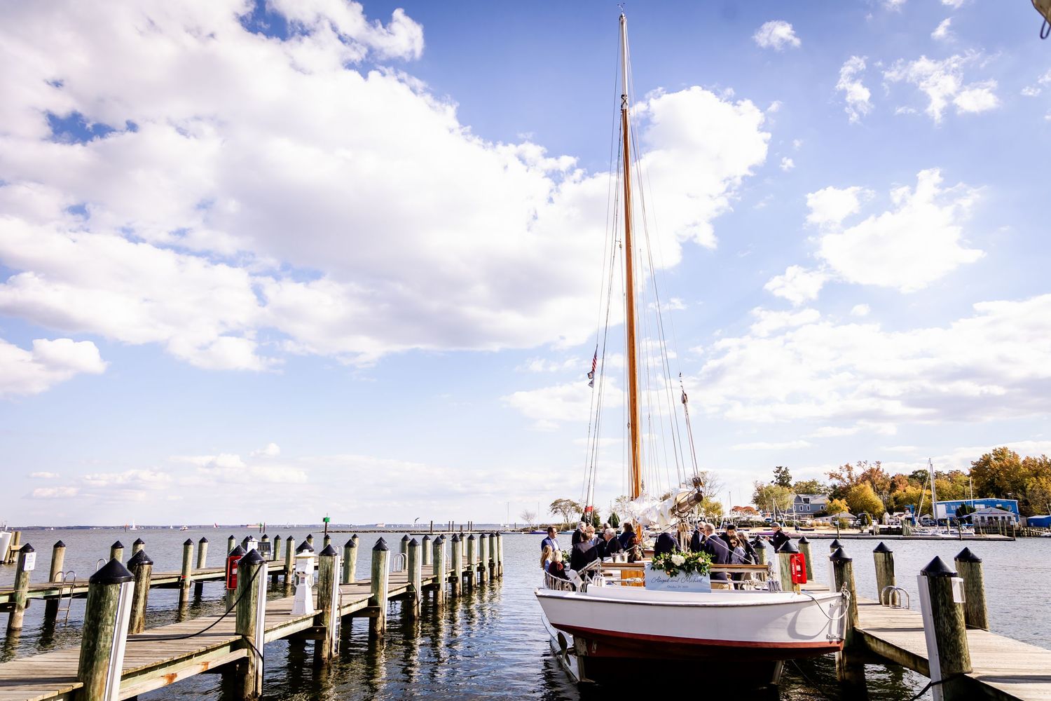 A sailboat docked at a wooden pier on calm waters under a partly cloudy blue sky at a waterfront venue.