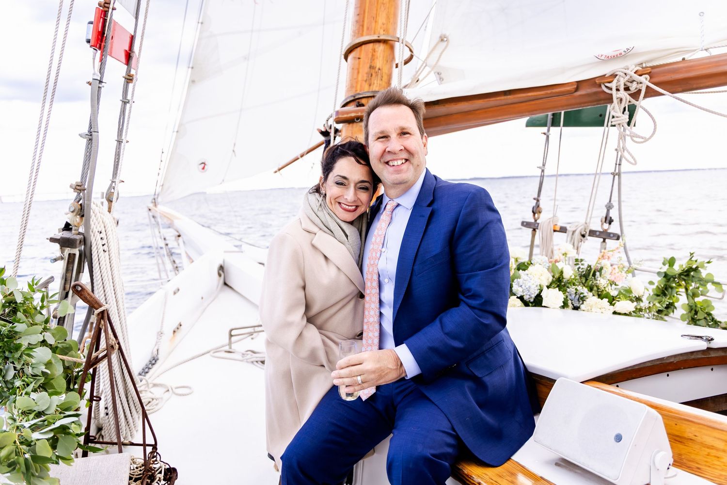 Couple smiling together on a decorated sailboat, man in blue suit and woman in beige coat on a sunny day.