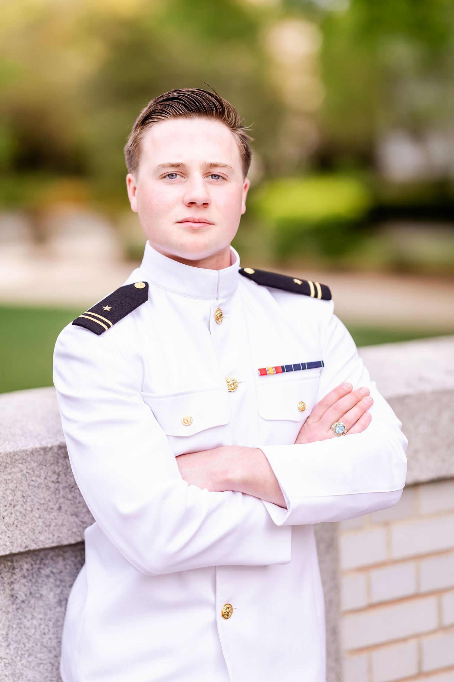 Naval officer in formal white uniform stands with crossed arms against stone wall.