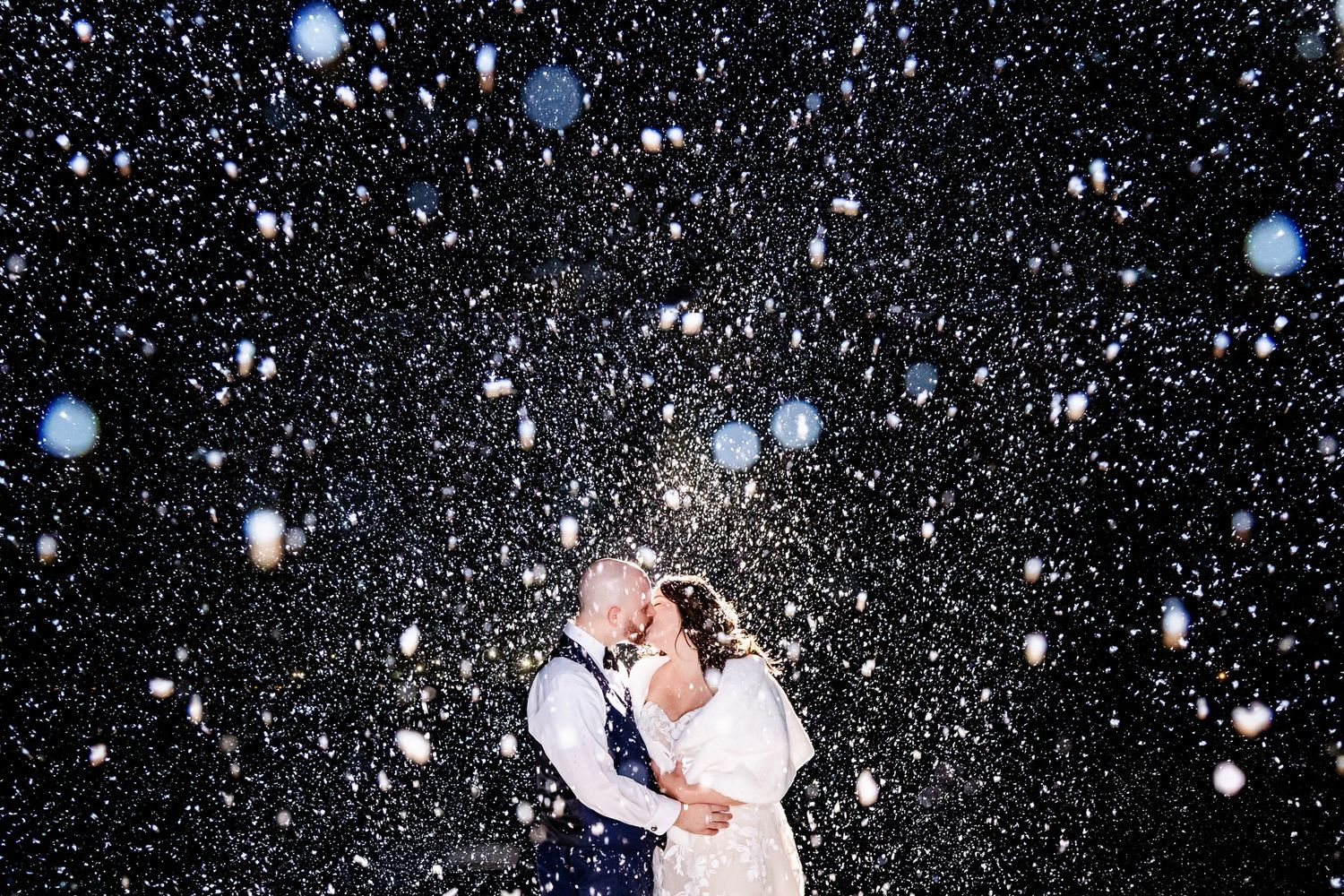 A romantic couple embraces under falling snow at night creating a magical winter wedding moment.