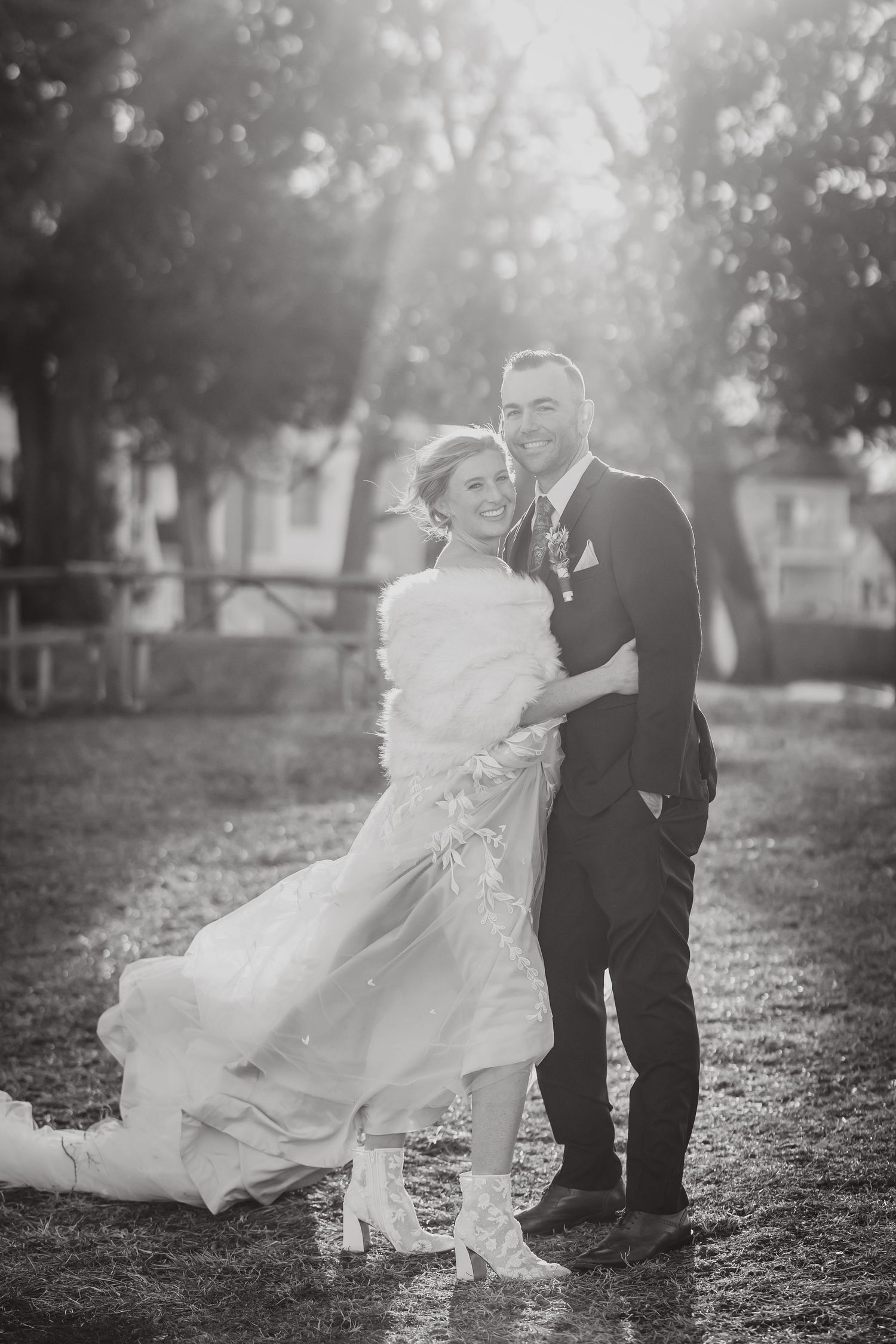 A romantic black and white wedding photo outdoors with sunlight streaming through trees in the background.