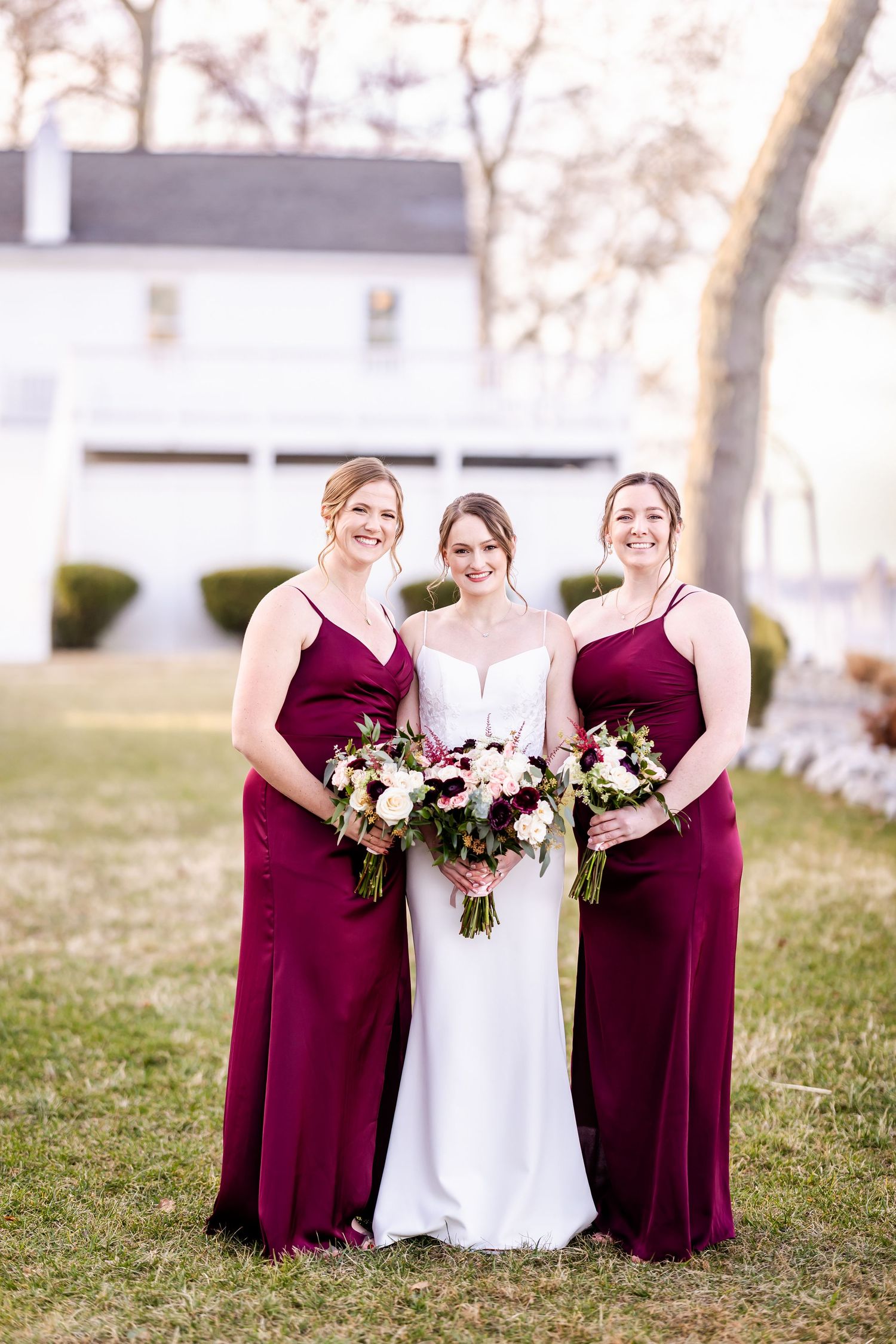 Wedding party in burgundy bridesmaid dresses and white bridal gown pose together on grassy lawn.