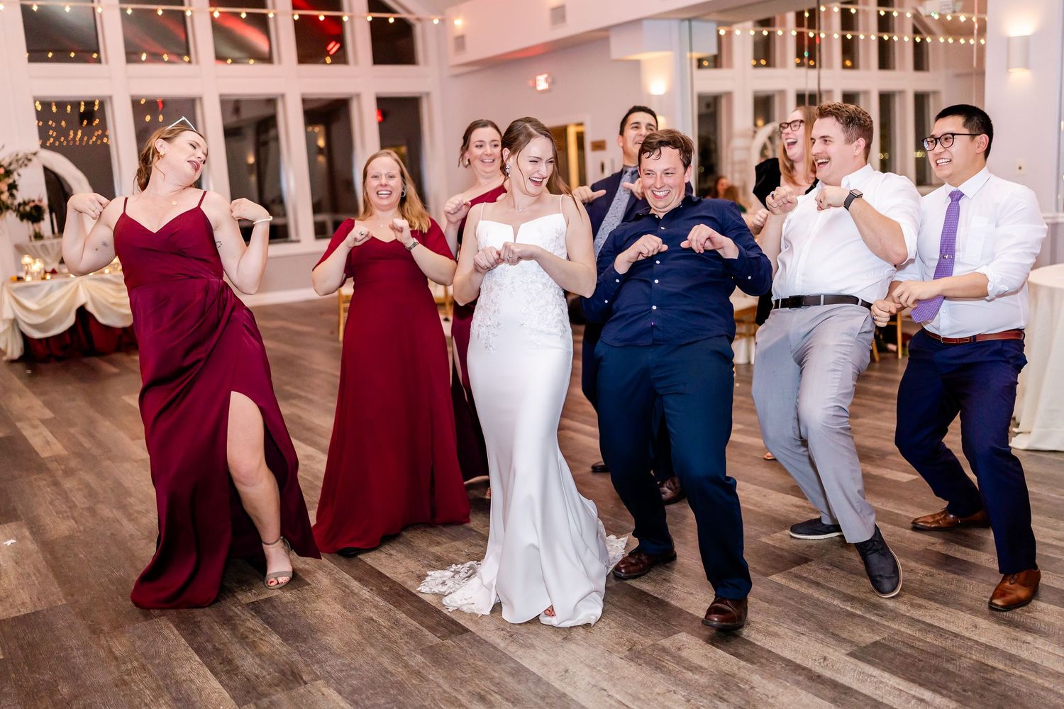 Wedding party poses together in navy and burgundy attire on wooden dance floor.