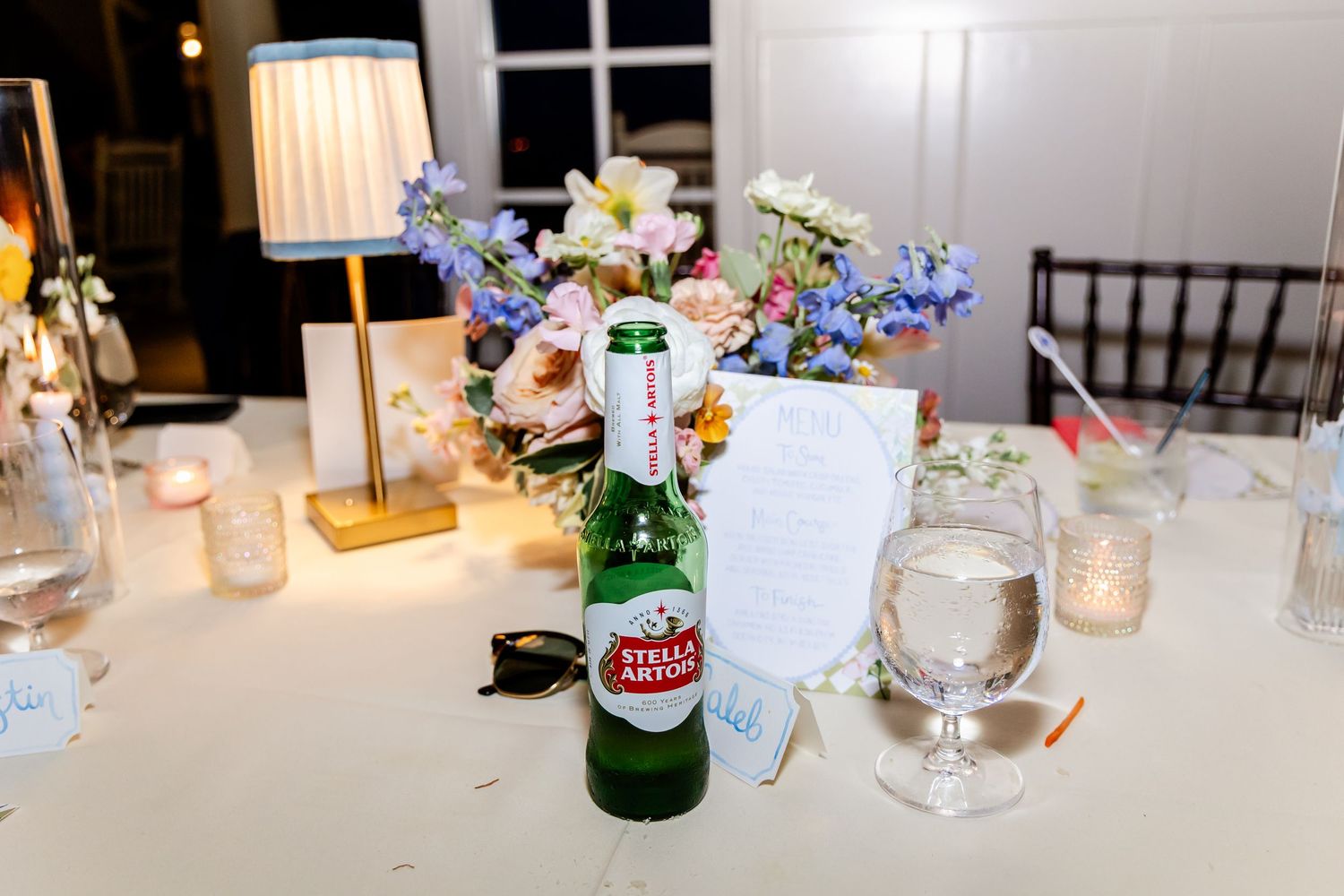 Wedding reception table setting with colorful floral centerpiece, candles, and a Stella Artois beer bottle.