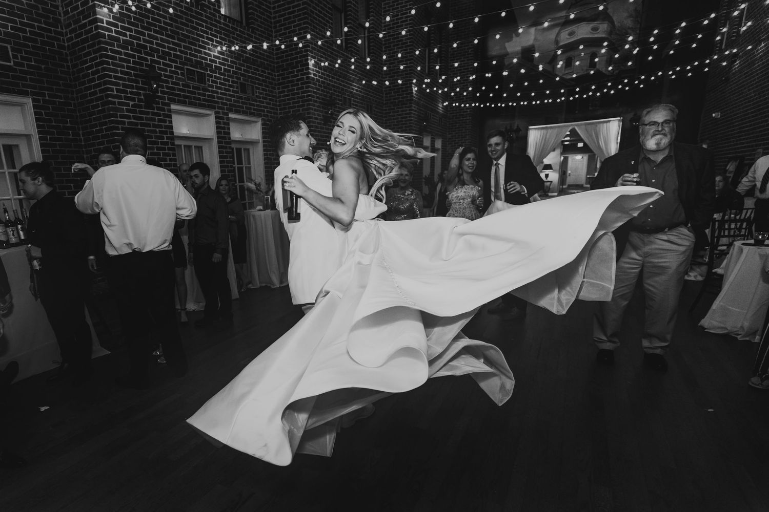 Black and white photo of a twirling couple dancing at an outdoor wedding reception under twinkling string lights.