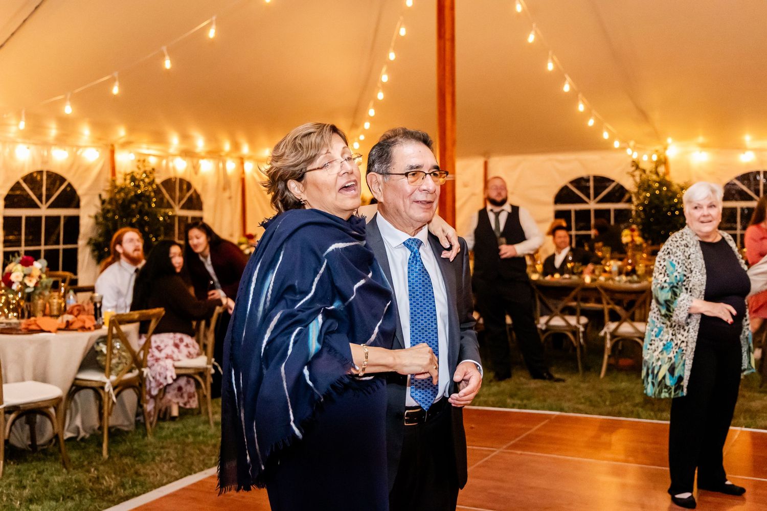 Wedding guests mingle and dance under string lights in an elegant white tent reception.
