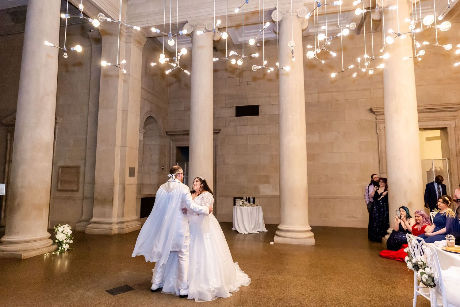 Wedding couple dances beneath twinkling lights in a classical columned ballroom.