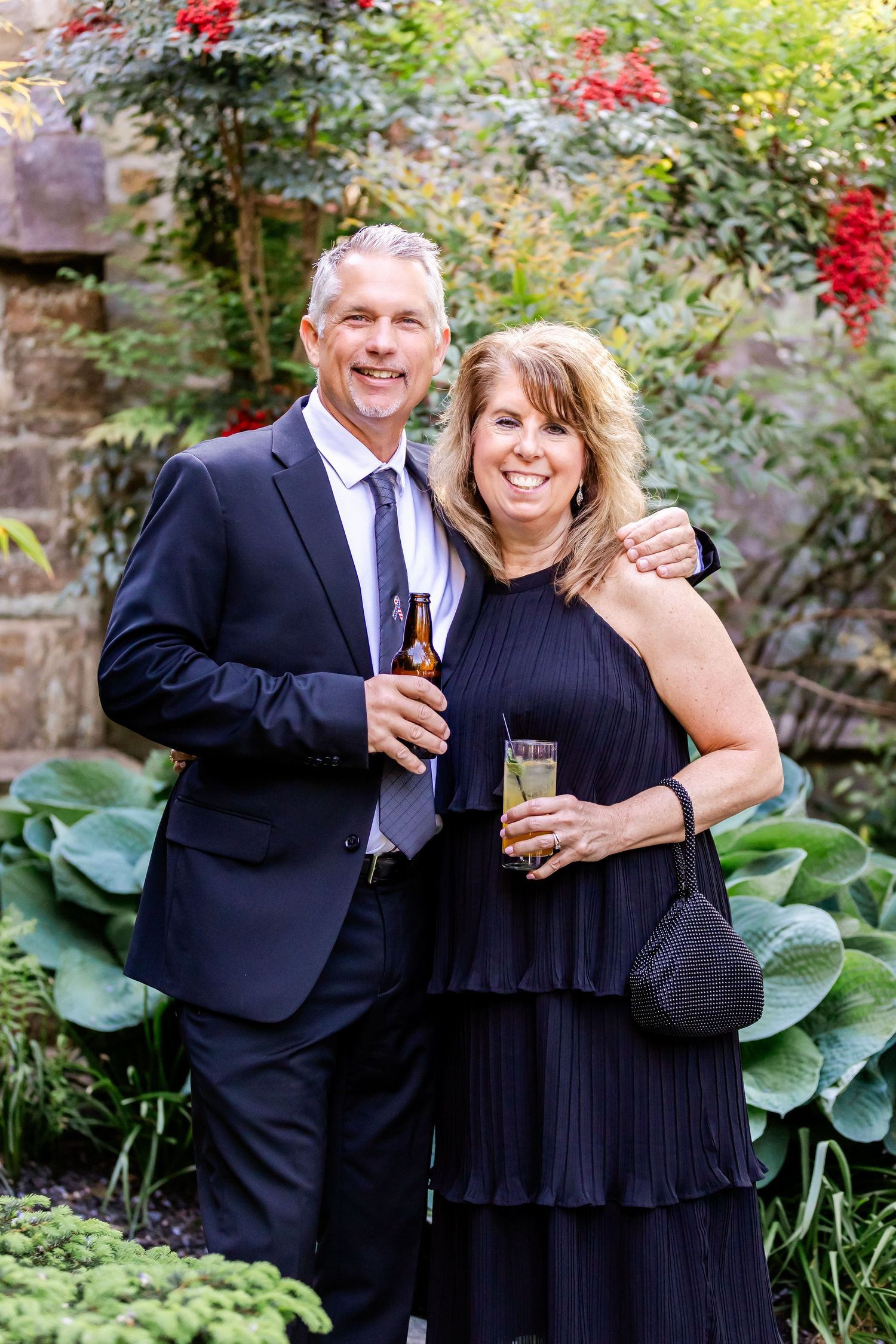 Couple in formal wear holding drinks in garden with red berries and lush foliage.