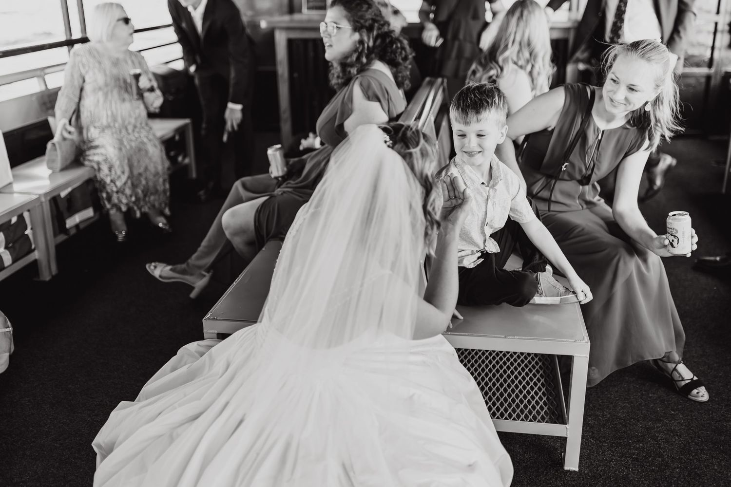 Black and white candid photo of a bride in white gown sitting with wedding guests during reception.