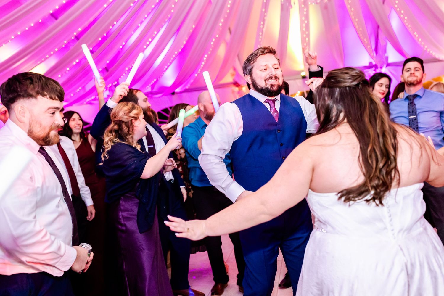 Wedding guests dancing and celebrating under a pink-lit tent with dramatic uplighting.
