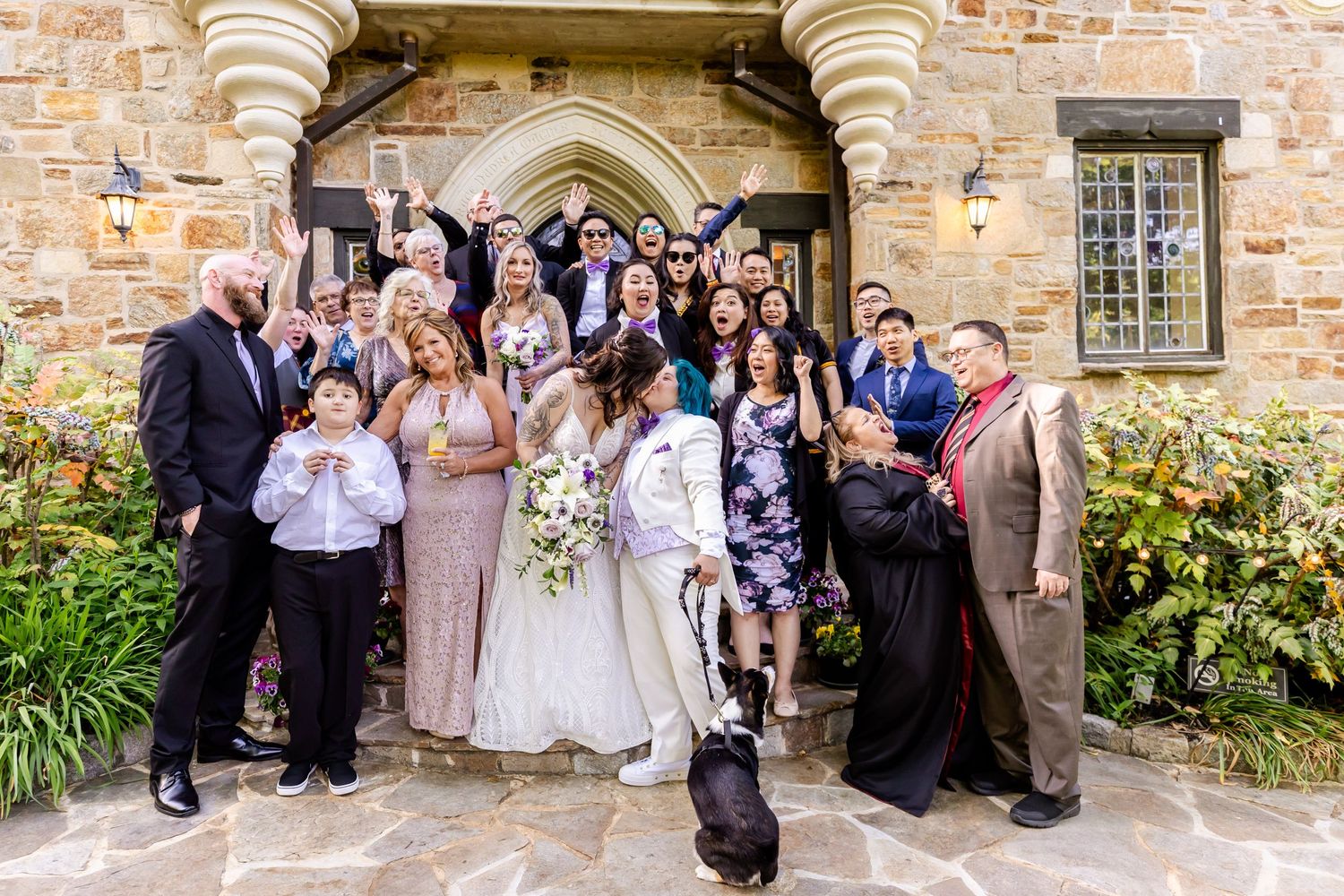 Large wedding group photo gathered in front of historic stone church entrance with decorative archway.