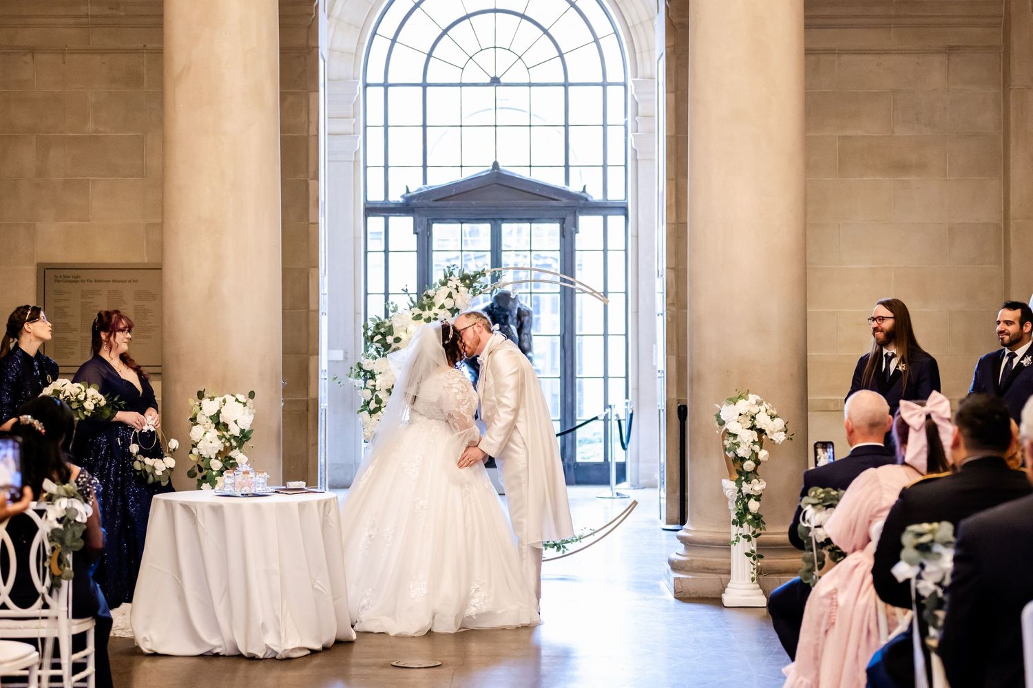 Wedding ceremony in an elegant venue with arched windows and floral decorations along the aisle.