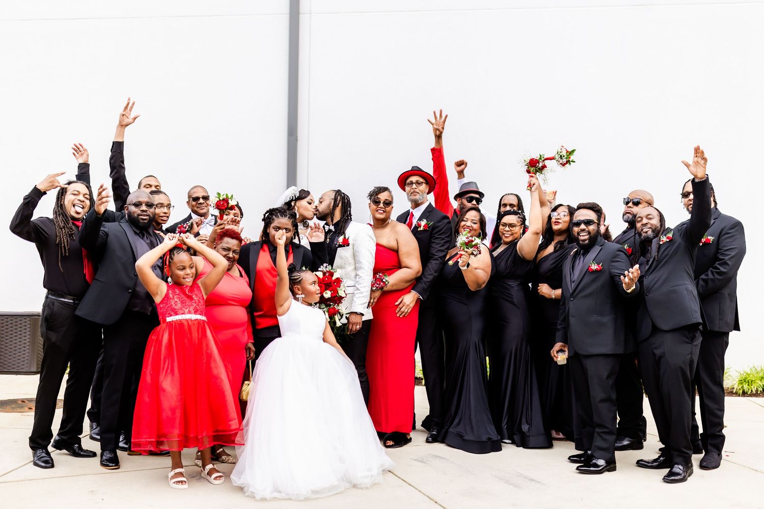 Large wedding party in red, black and white attire celebrate with raised arms against wall.