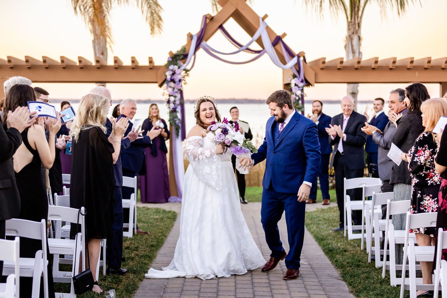 Newlyweds celebrate during recessional walk down outdoor aisle under palm trees with guests cheering.