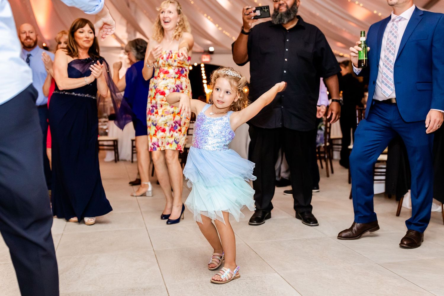 A small child in a light blue dress dances enthusiastically at a formal event while others watch from the sidelines.