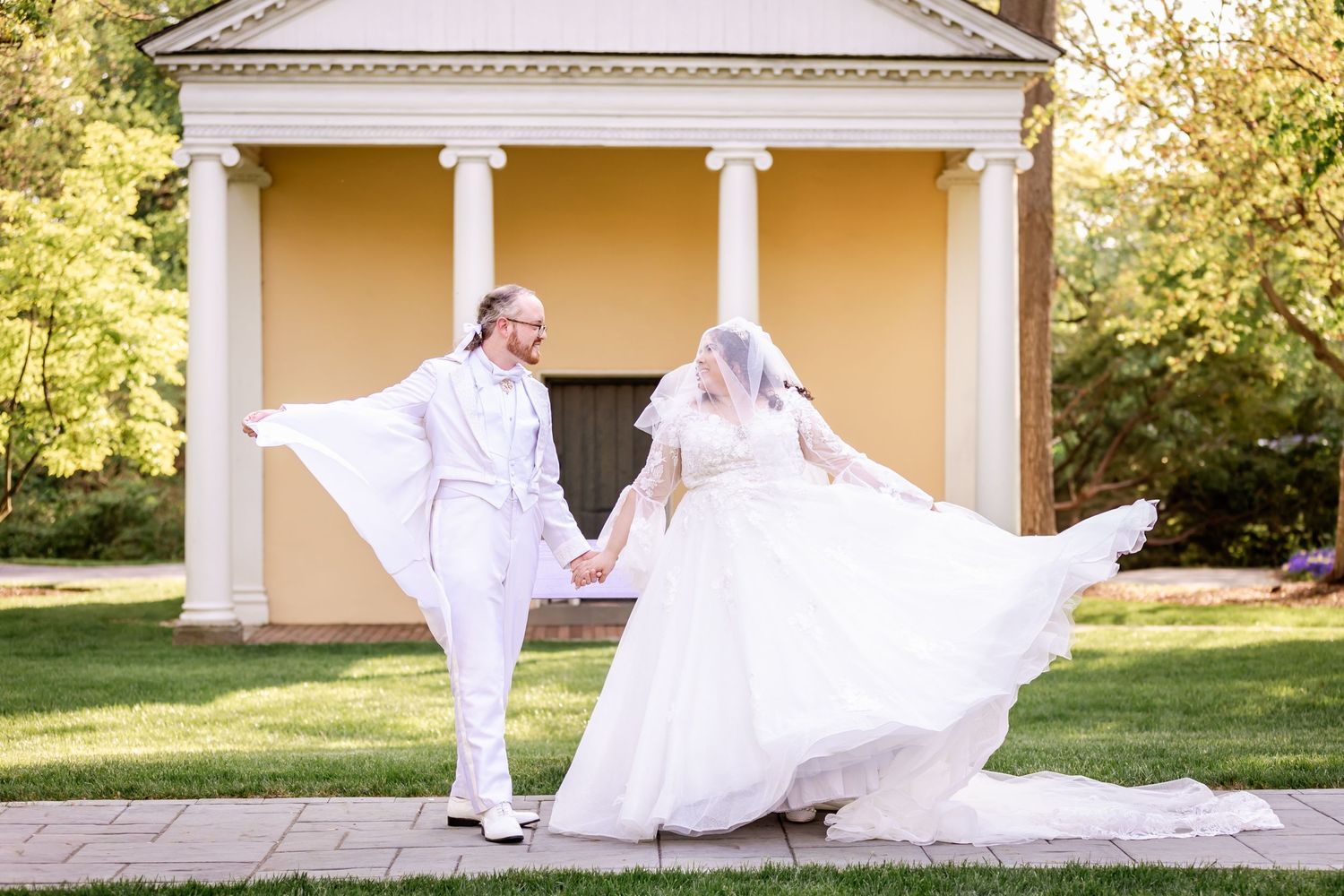Elegant wedding couple twirls playfully in front of classic yellow colonial building.