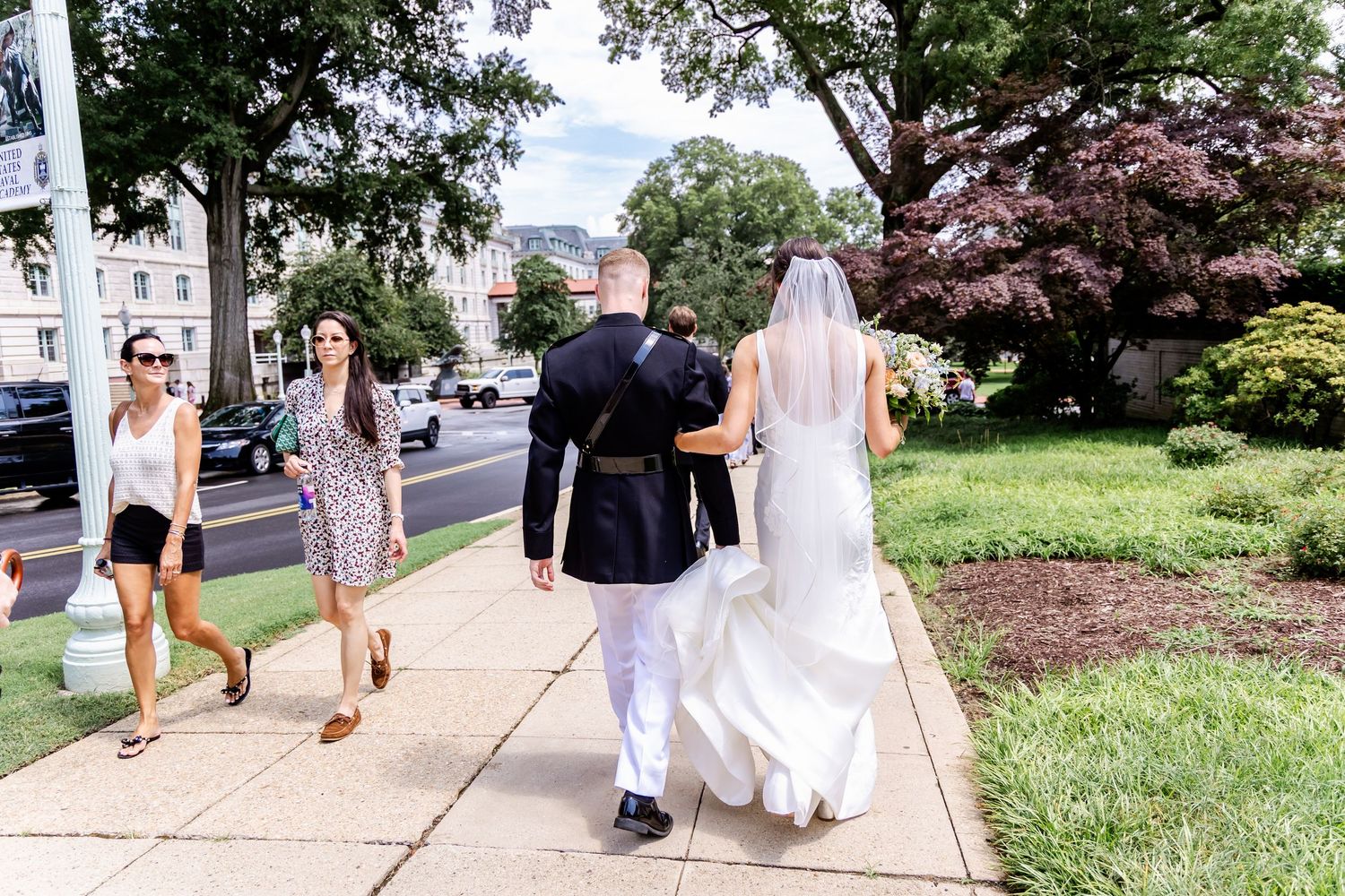 A bride in white dress walks with others along a city sidewalk on a sunny summer day.