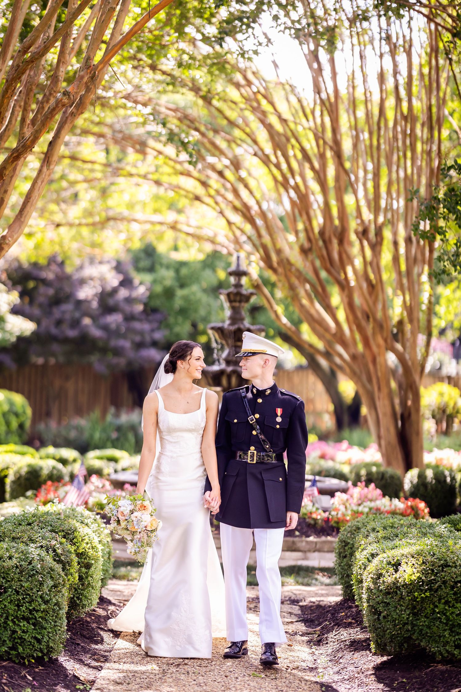 A bride in white dress walks with military groom under an archway of blooming trees.