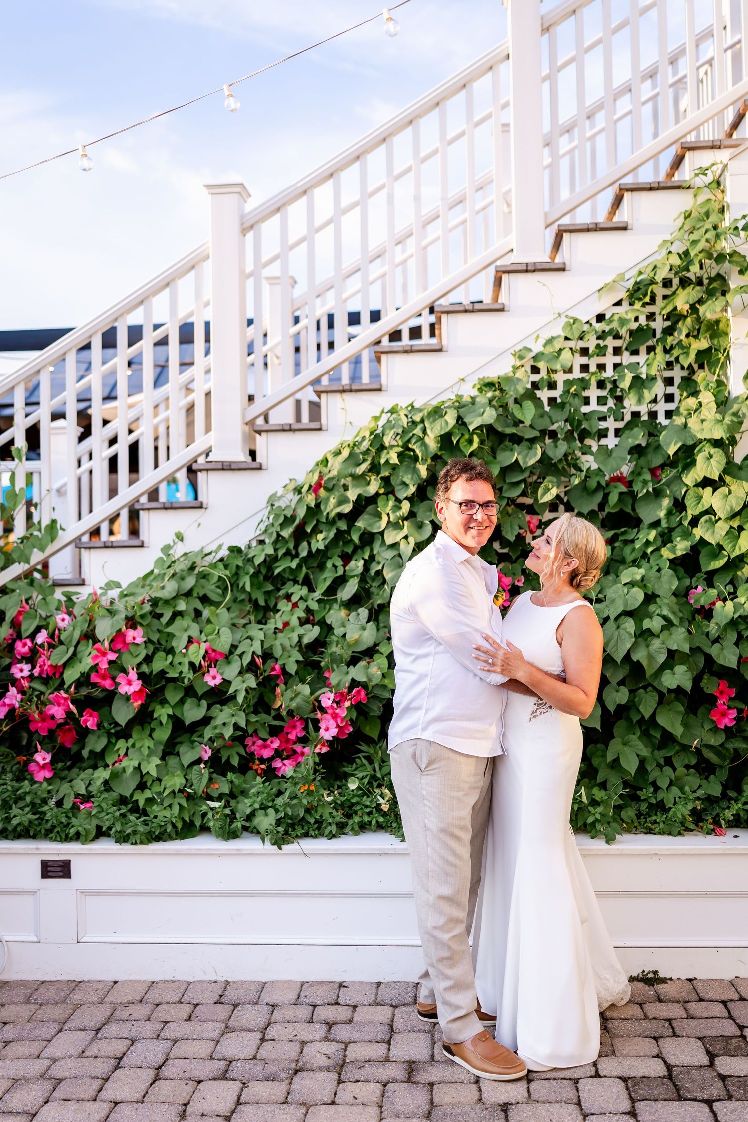 Romantic couple shares first dance on brick patio with flowering vine wall and white staircase in background.
