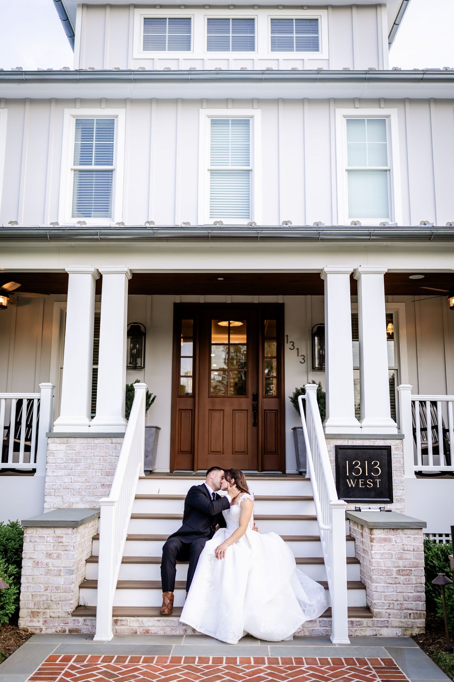 Romantic wedding couple embraces on white farmhouse porch with stone accents and wooden columns.
