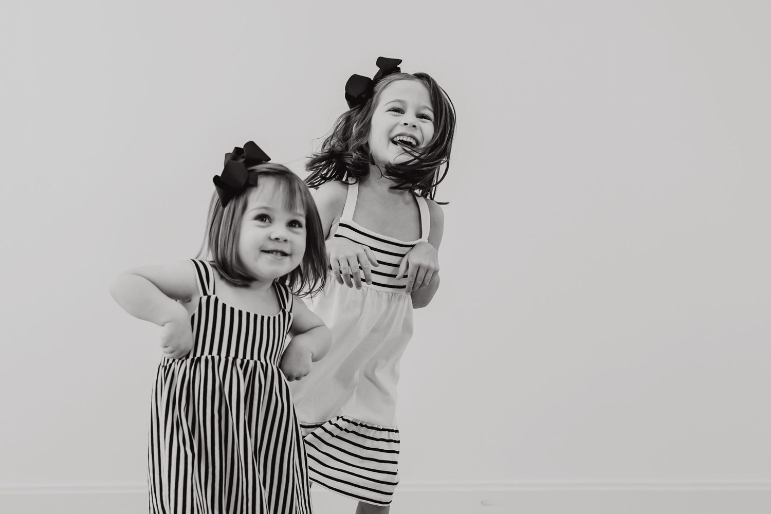 Two children in striped dresses pose together against a white wall in black and white photography.