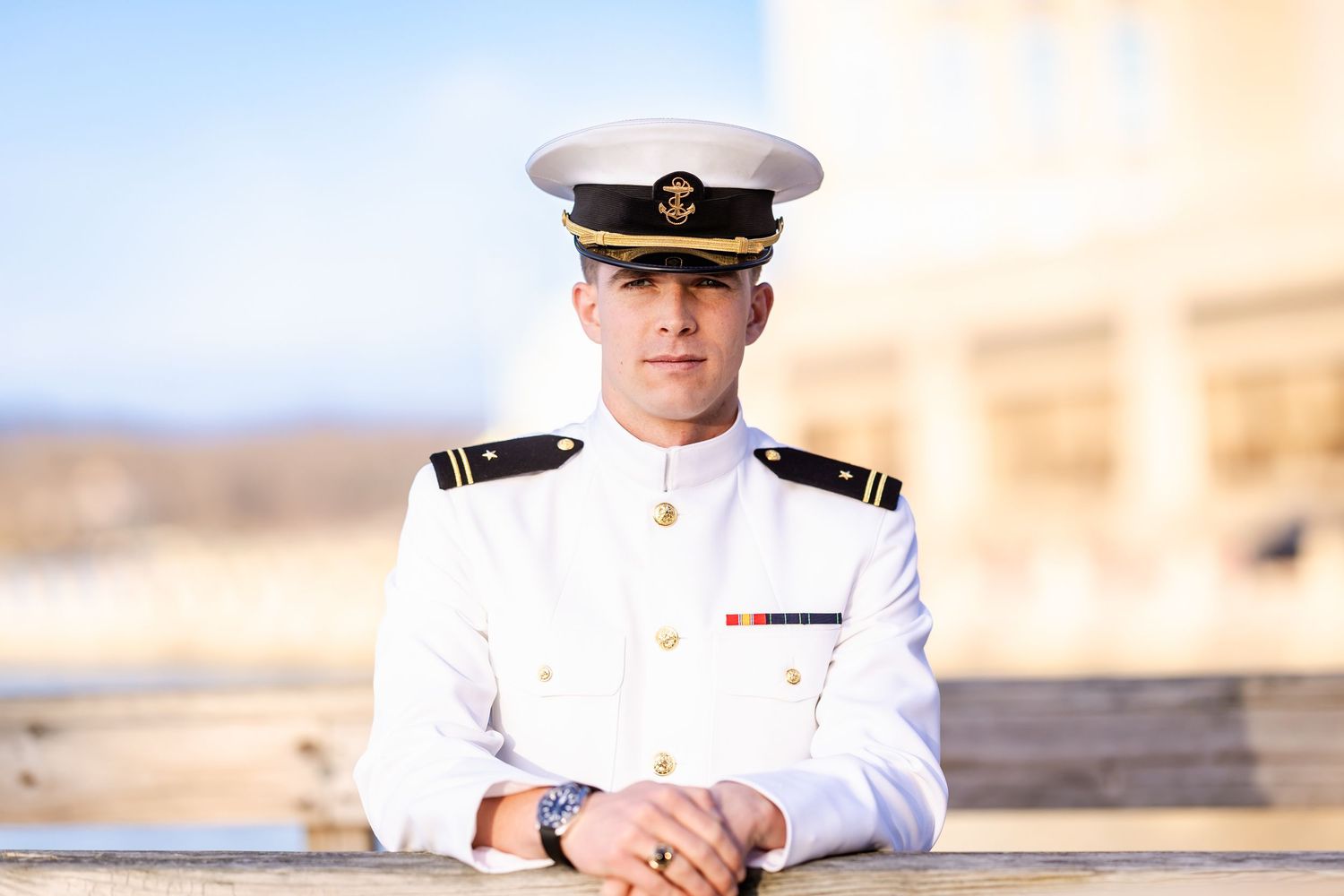 Young naval officer in white dress uniform and cap poses confidently outdoors in bright sunlight.