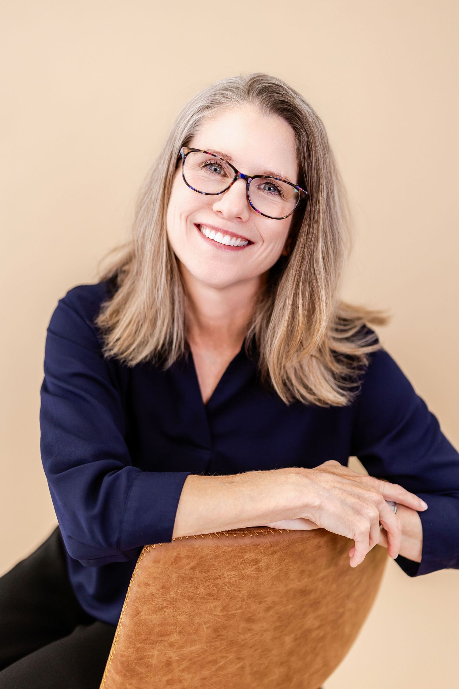 Smiling middle-aged woman with glasses and navy blouse seated casually against a beige background.