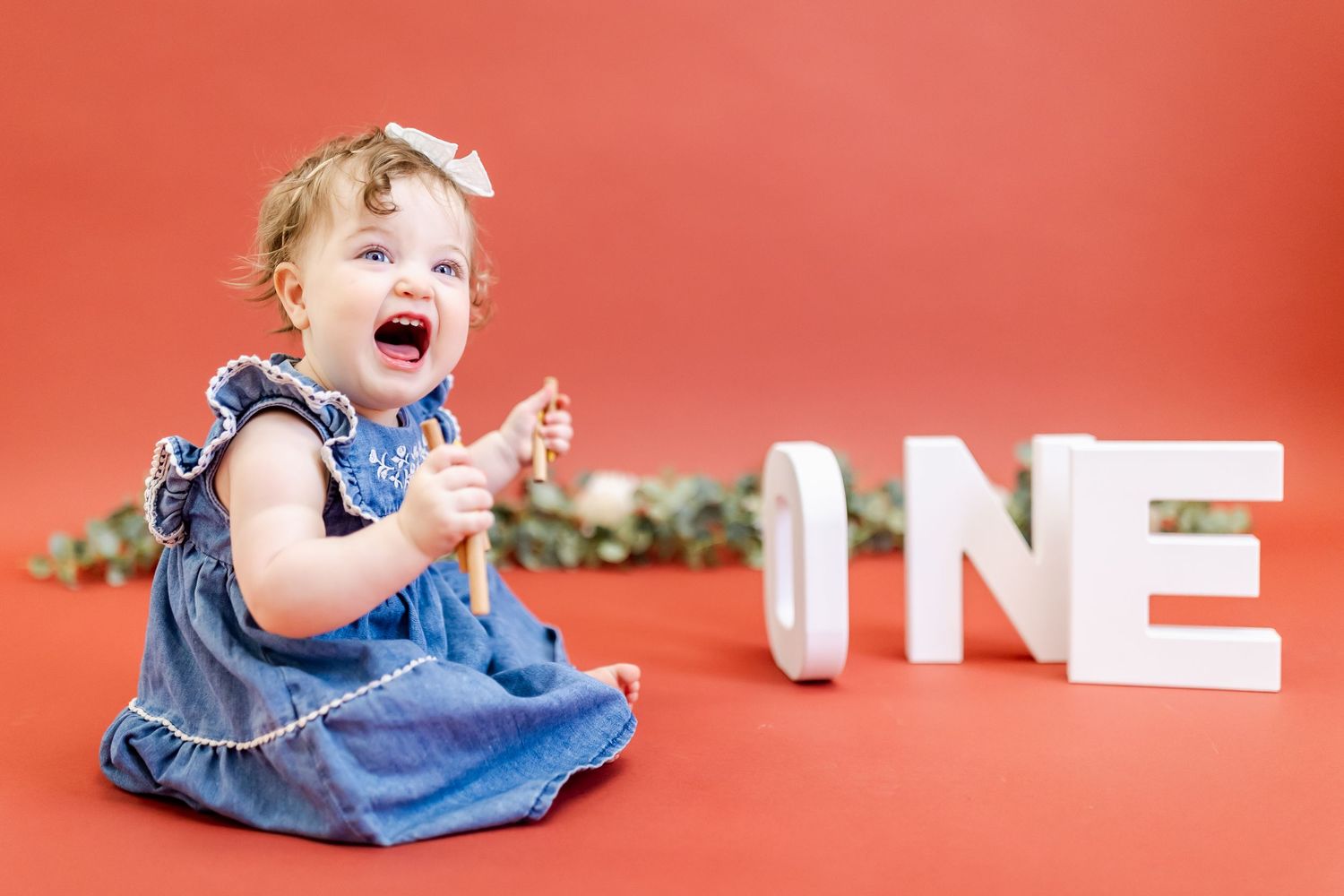 A joyful baby in a blue dress sits on a red backdrop next to white 'ONE' letters for a first birthday photoshoot.