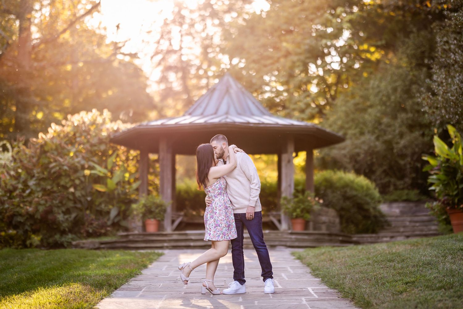 A couple embraces in front of a garden gazebo during golden hour sunset in a romantic outdoor setting.