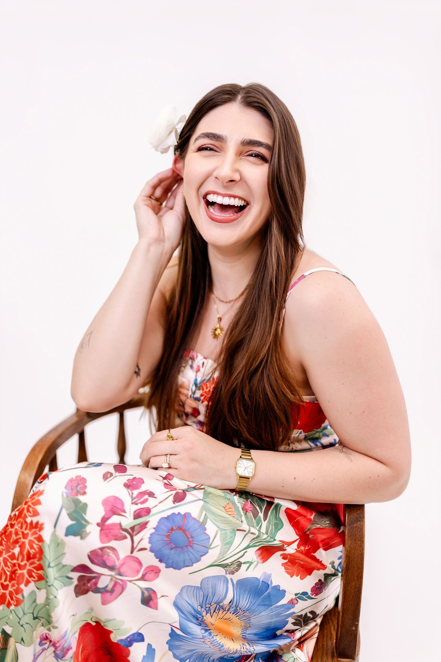 Happy woman in floral dress with flower in hair laughing while seated in wooden chair against white background.