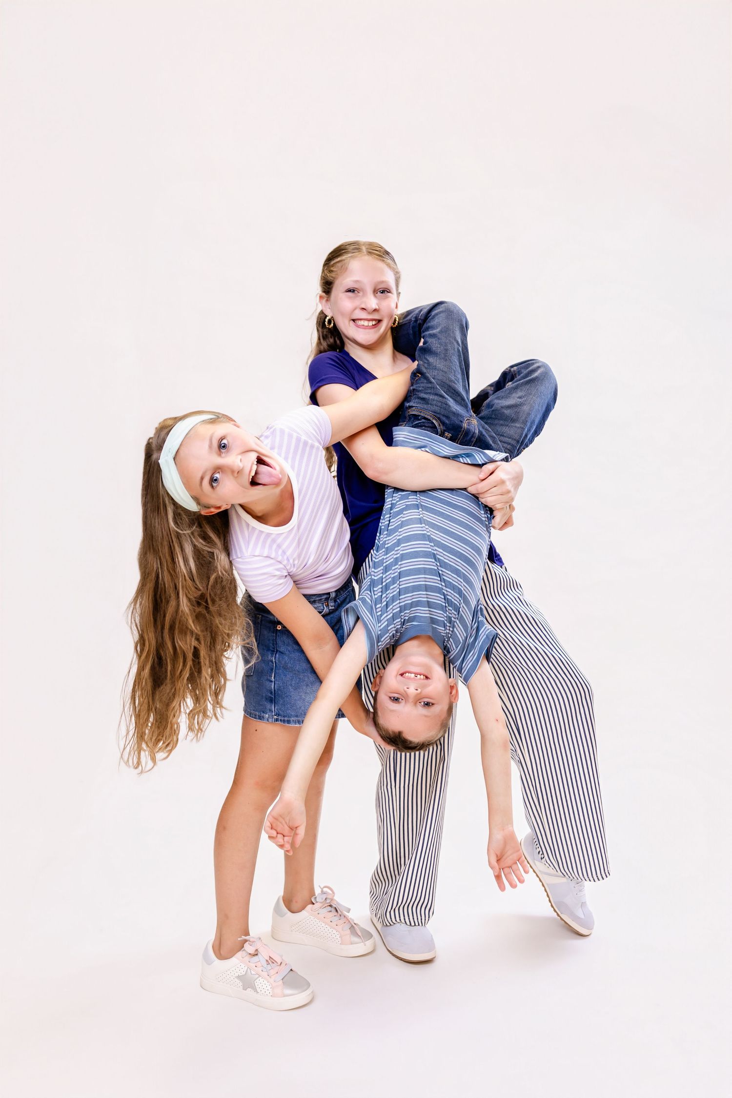 Three playful kids posing together, one held upside down, laughing and having fun against a white background.