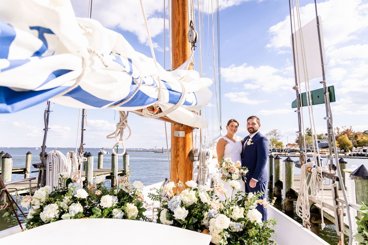 Couple poses on decorated sailboat deck with white floral arrangements, blue sails, and waterfront marina backdrop on wedding day.