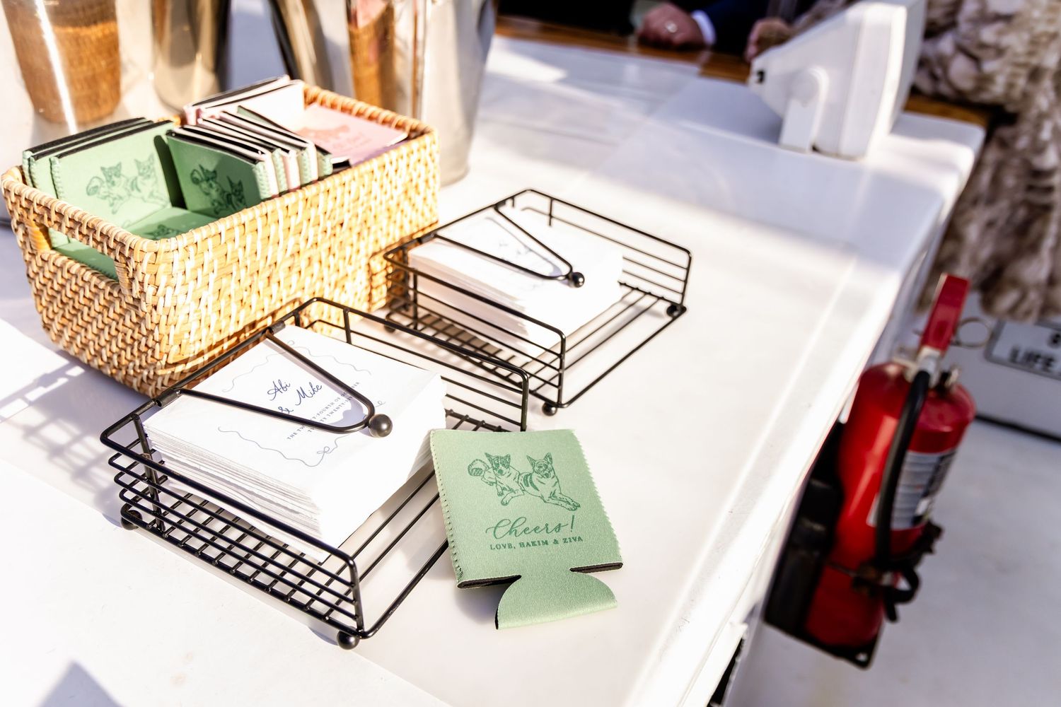 Wire rack organizers and wicker basket holding custom green wedding koozies and cards on a white table.