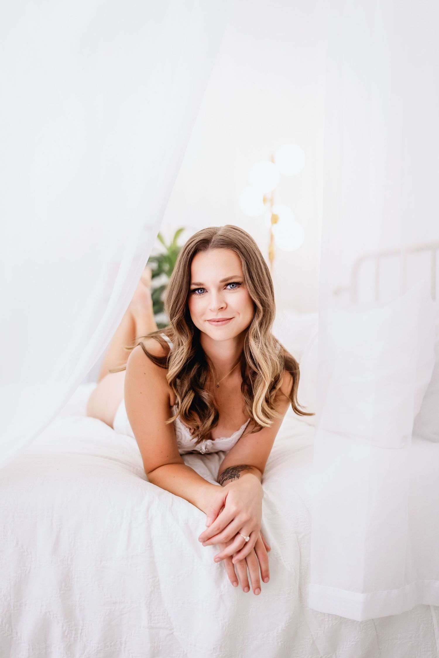 Portrait of someone relaxing on white bedding with wavy brown hair and wearing a neutral-colored top.