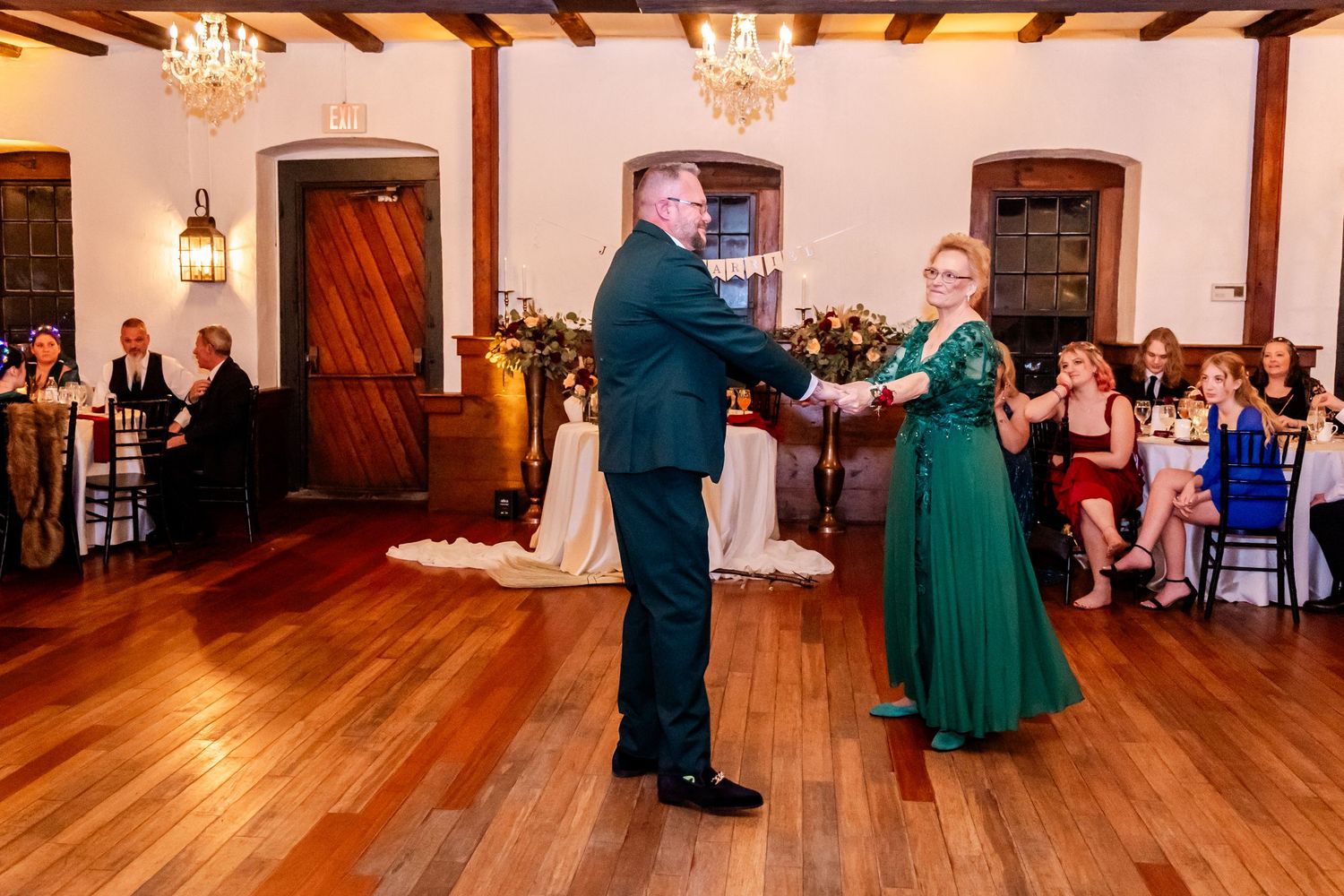 Couple dancing together in an elegant ballroom with wooden floors and exposed beams while guests watch from tables.