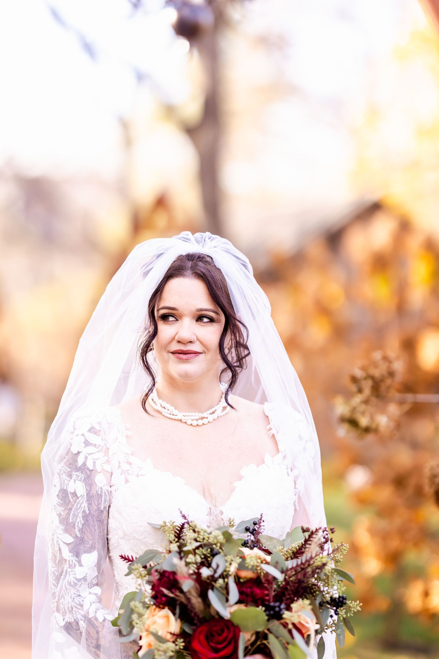 A bride in a lace wedding dress holds a burgundy and cream bouquet against an autumn backdrop.