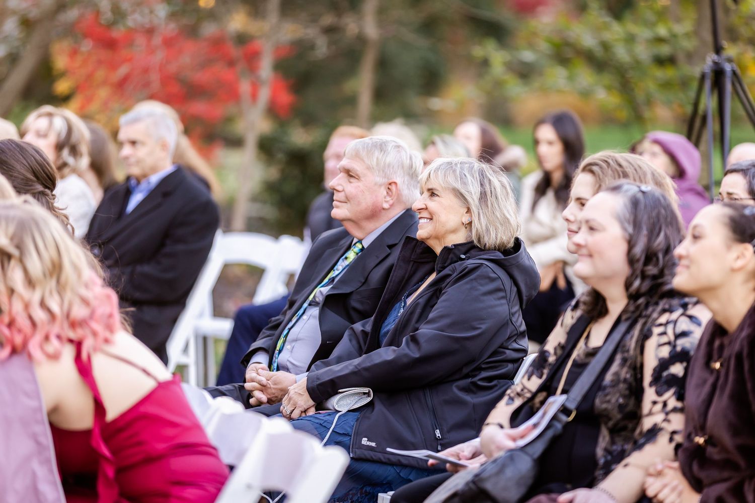 Wedding guests seated outdoors watching ceremony with fall foliage in background.
