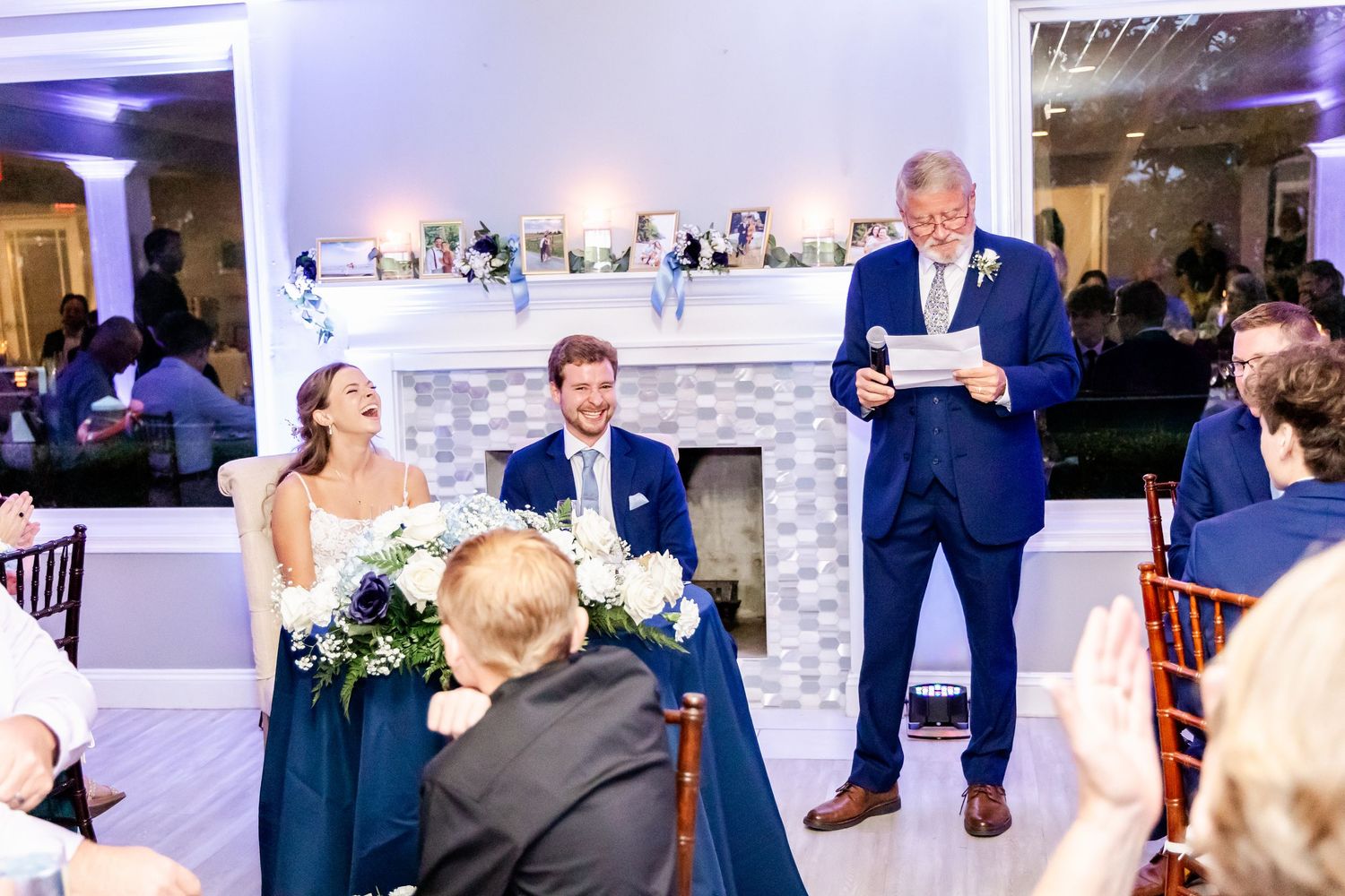 Wedding guests listen to a speech during reception at elegant white venue with fireplace mantle.